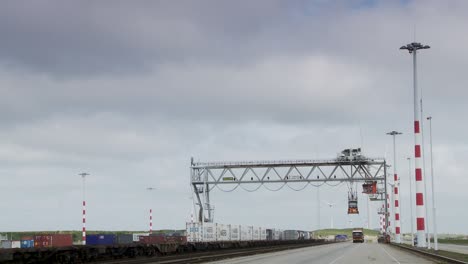 Tren-De-Carga-Pasando-Bajo-Una-Grúa-Pórtico-En-Una-Terminal-De-Contenedores,-Cielo-Nublado