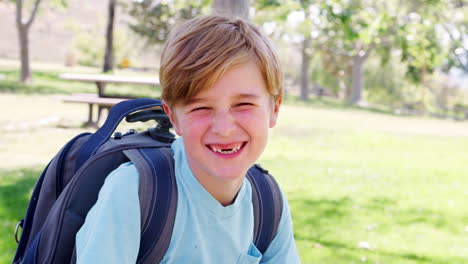 slow motion portrait of young boy with backpack in park smiling at camera