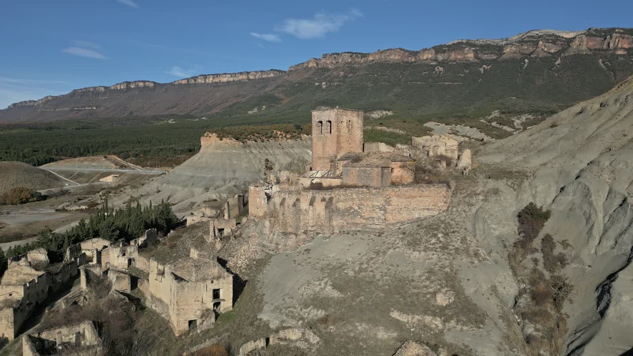 Vista aerea con drone che rivela gli edifici in pietra in rovina e la chiesa della storica città fantasma di Esco, immersa nelle aride colline dell'Aragona, Spagna, sotto un cielo azzurro e limpido.
