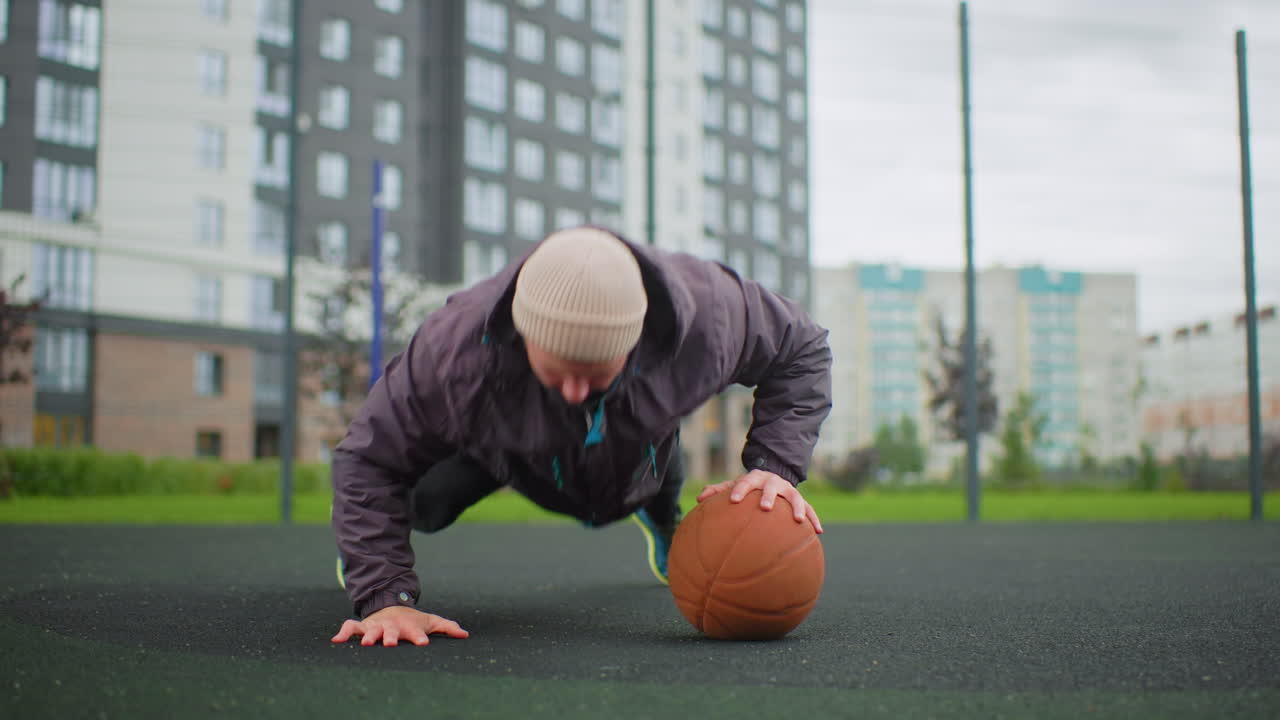 man balancing on basketball outdoor court, performing dynamic pushup progression, wearing beanie and jacket, overcast sky, city buildings in background, rubber asphalt surface, focused expression