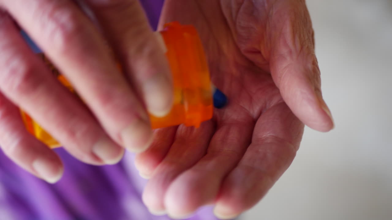 An elderly woman pouring pills from a prescription medicine bottle into her wrinkled, aged hand to cure her illness