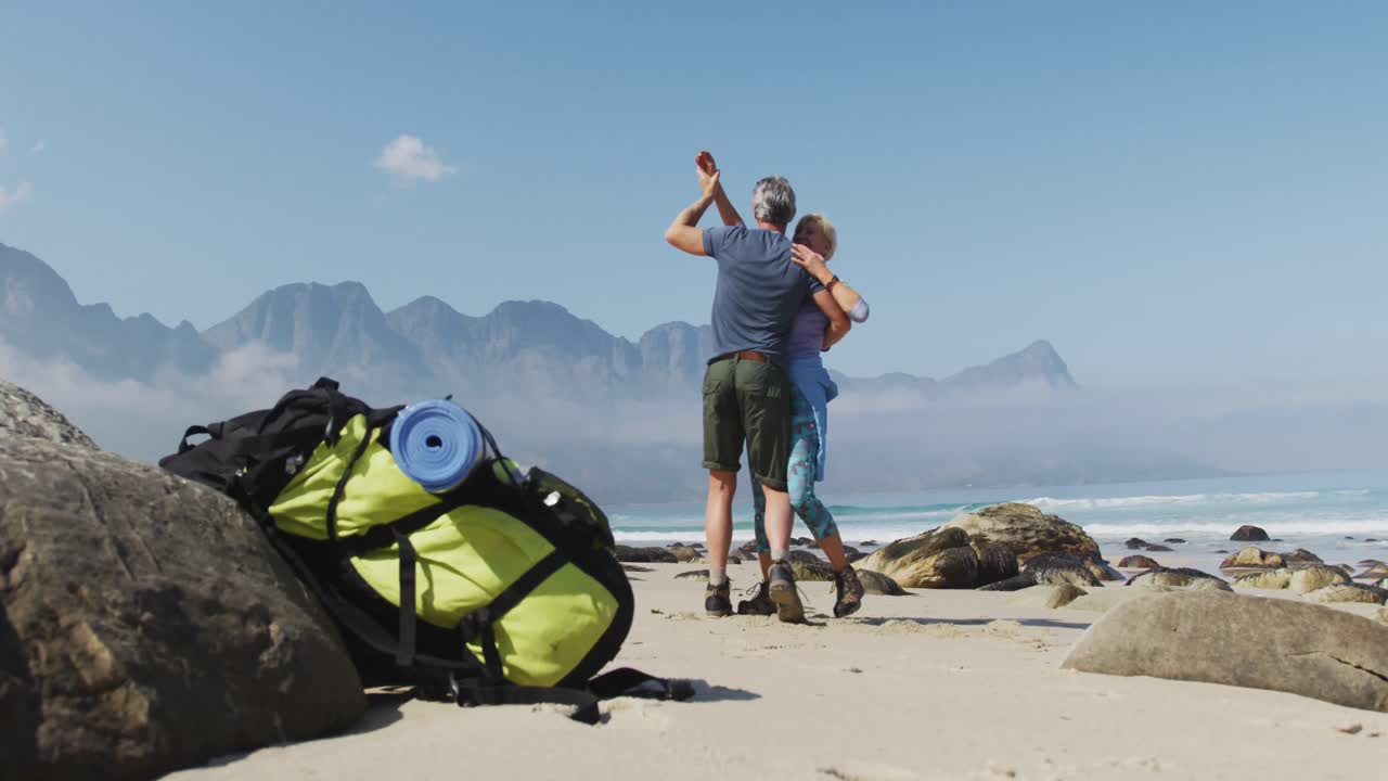 una pareja de excursionistas senior bailando en la playa mientras caminan.