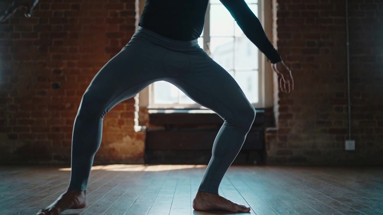 Ballerina che si abbassa, accovacciata e si muove lateralmente in un loft vicino a una finestra alta, indossando top e leggings