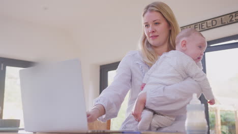 working mother using laptop at home whilst cuddling baby son over shoulder- shot in slow motion