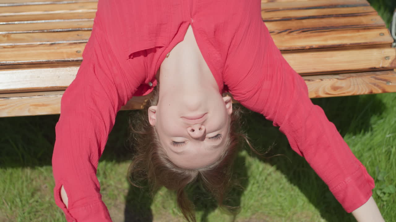 Young woman in red top lies on wooden swing bench with hands raised to block direct sunlight from hitting her face, bright natural light casting shadows across her features