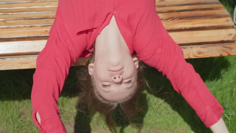 young woman in red top lies on wooden swing bench with hands raised to block direct sunlight from hitting her face, bright natural light casting shadows across her features