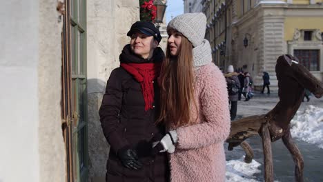 Two-smiling-women-tourists-looking-at-shop-window-on-city-street,-family-couple-talking,-embracing