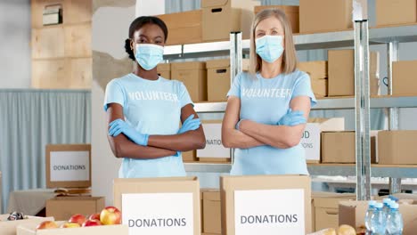 african american and caucasian women volunteers in facial masks packing donation boxes and looking at camera in charity warehouse