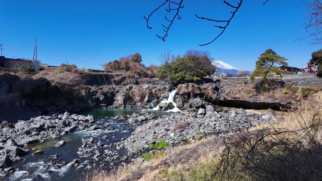 Küçük bir şelalesi olan, bir şehirden akan kayalık bir nehir ve ufukta Fuji Dağı'nın büyüleyici manzarası