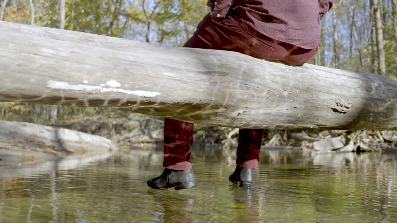 Ripresa da dietro: Uomo con vestiti bordeaux siede su un tronco sopra il fiume. Carrellata, concetto di solitudine.