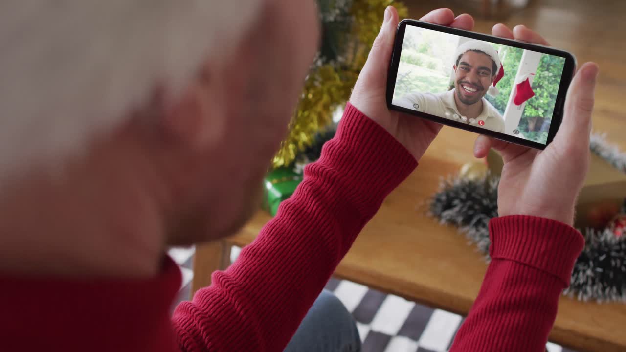 hombre caucásico con sombrero de santa usando un teléfono inteligente para una videollamada de navidad con un hombre sonriente en la pantalla
