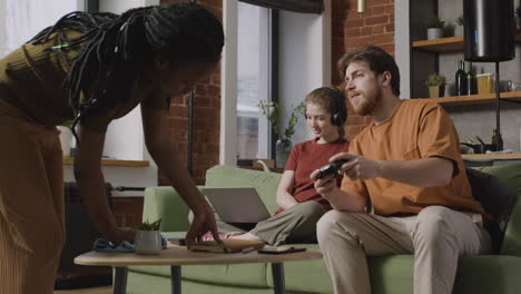 chica usando laptop y chico jugando videojuegos sentados en el sofá mientras su compañera de cuarto limpia la casa 1