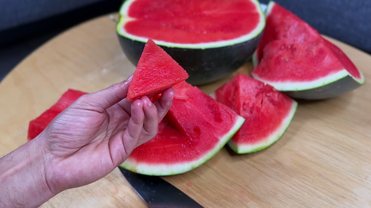 Close-up of a hand holding a triangular slice of juicy watermelon with more pieces in the background, perfect for food, health, summer, lifestyle, and nutrition projects