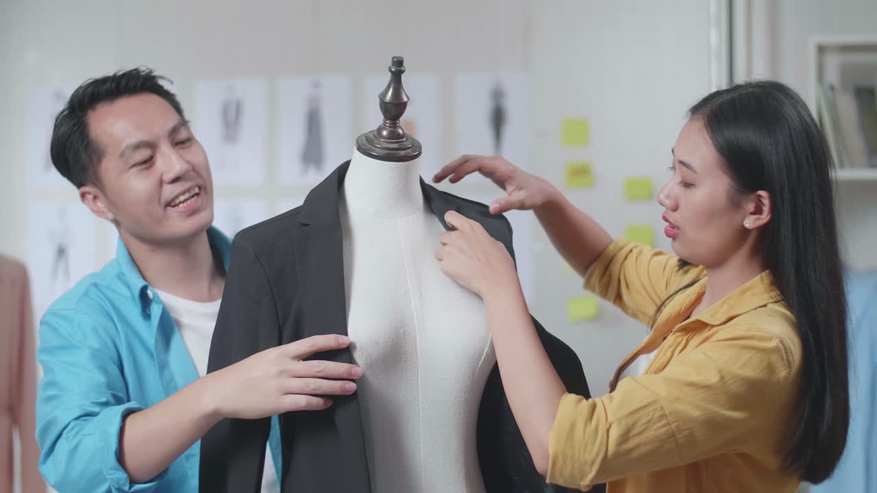아시아의 남녀 디자이너들이 바느질 기계와 함께 스튜디오에서 옷을 디자인하는 동안 마네킨에 비즈니스 재을 입고 서로를 돕고 있습니다.
