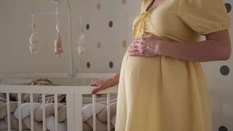 pregnant woman in yellow dress rubbing her belly with left hand while standing next to white wooden crib in nursery with dotted wall in the background