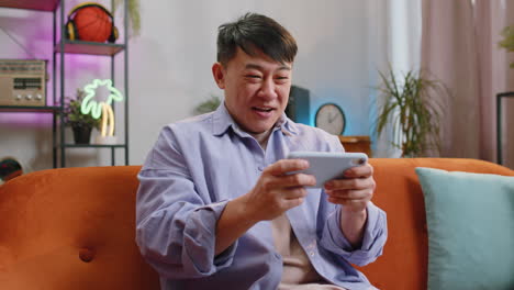 Worried-asian-handsome-man-playing-racing-or-shooter-video-games-on-smartphone-at-home-on-couch