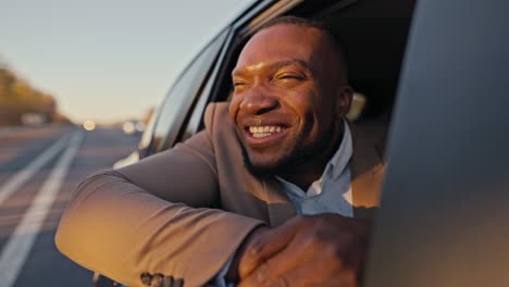 Happy-cheerful-male-businessman-with-Black-skin-in-a-brown-suit-smiles-broadly-and-looks-out-of-the-rear-window-of-a-modern-car-during-his-trip-outside-the-city