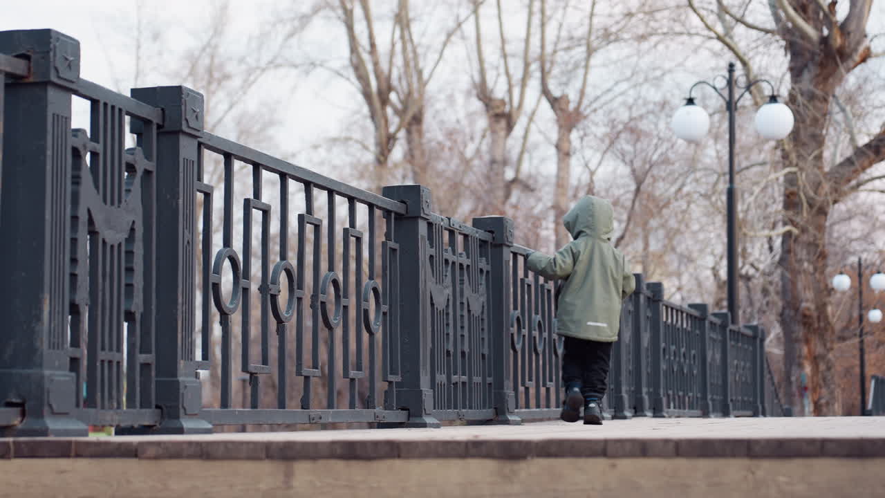 Young boy in green winter jacket and boots walking playfully on elevated pedestrian bridge with black iron railings in serene winter park filled with leafless trees and lampposts under cloudy sky