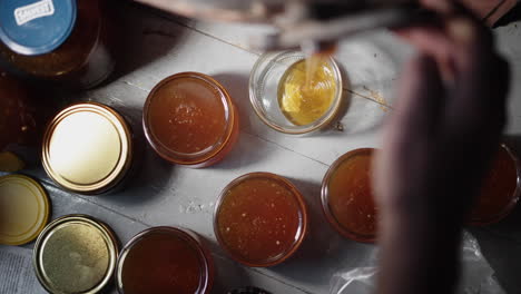 Top-Down-Slow-Motion-View-of-Pure-Honey-Glass-Jars,-Farmer-Hand-Holding-Touching-the-Bee-Honey-Extractor-Faucet-while-it's-Flowing-and-Dripping-into-the-Bottle