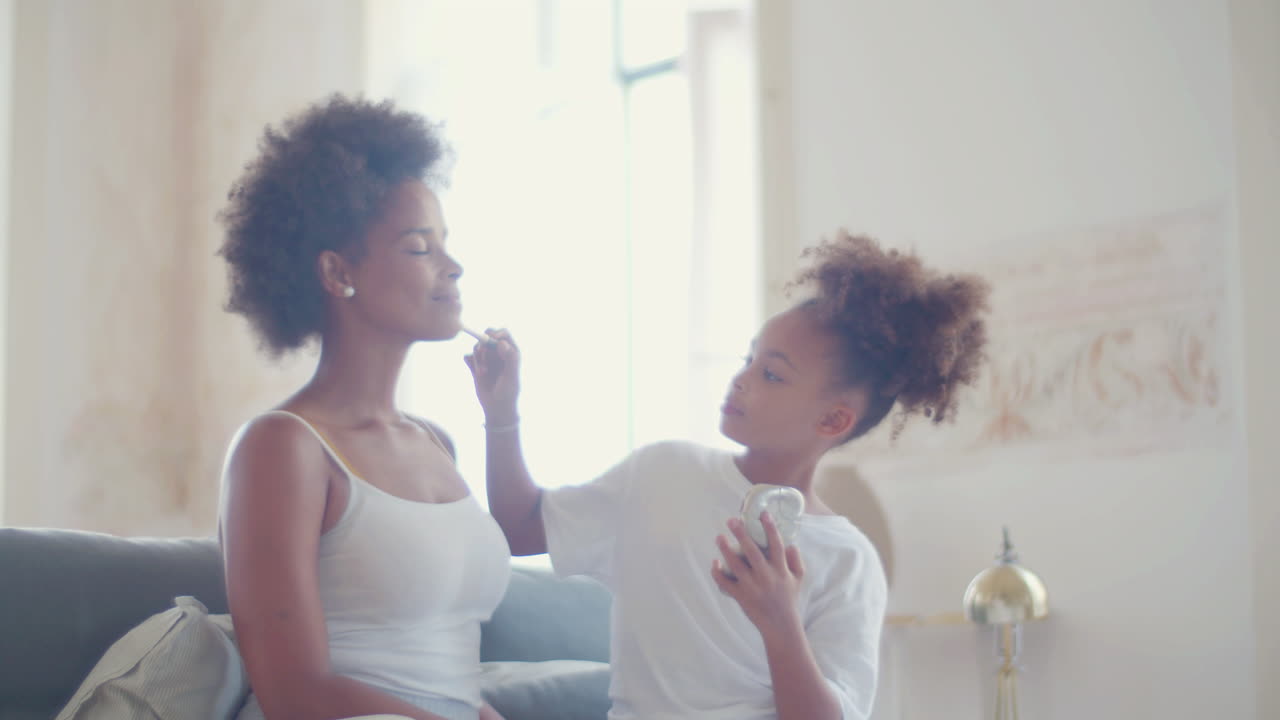 Cute girl doing make-up on her mother sitting on couch at home