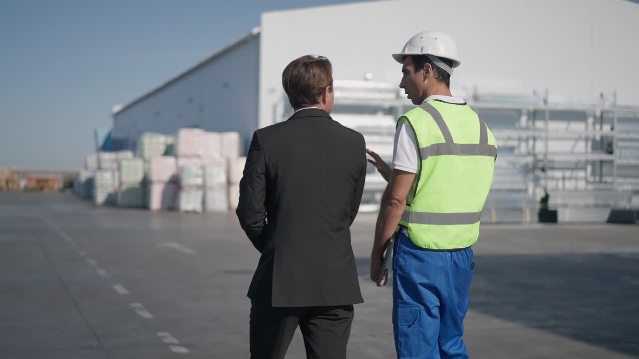 concentrado hombre confiado de oriente medio en uniforme y gerente de almacén caucásico en traje caminando en cámara lenta hablando. la cámara en vivo sigue al empleado y el empleador discutiendo negocios al aire libre.