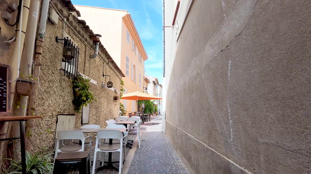 Una carrellata dal basso percorre uno stretto vicolo mediterraneo oltre i tavoli vuoti di un ristorante.