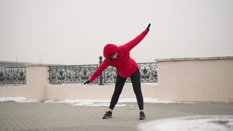 woman in red hoodie and gloves performing side stretch outdoors on snow-covered path near decorative metal fence with blurred view of distant bridge in foggy winter atmosphere