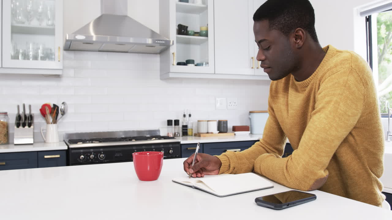 Using smartphone and notebook, African American man in kitchen preparing for holidays, copy space
