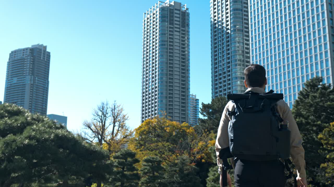 A man walks along a scenic path in Hamarikyu Gardens, Tokyo, surrounded by vibrant autumn foliage in shades of red, orange, and yellow.