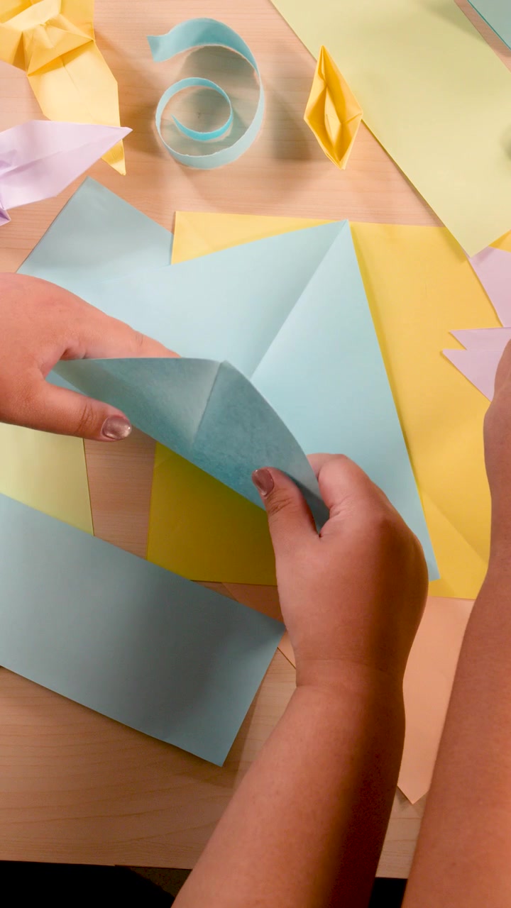 Hands carefully fold blue paper on desk with colorful origami, bright lighting, overhead camera angle