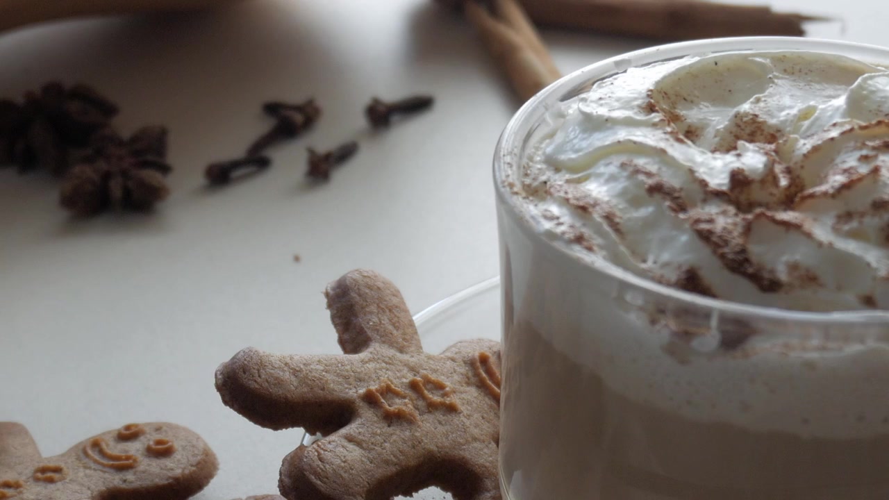 Zoom su una tazza di vetro trasparente con caffè o cacao con panna, cannella, biscotti di Natale a forma di omino di pan di zenzero e chiodi di garofano. Sapori natalizi.