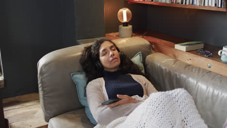 biracial woman lying on sofa under blanket listening music using smartphone at home, slow motion