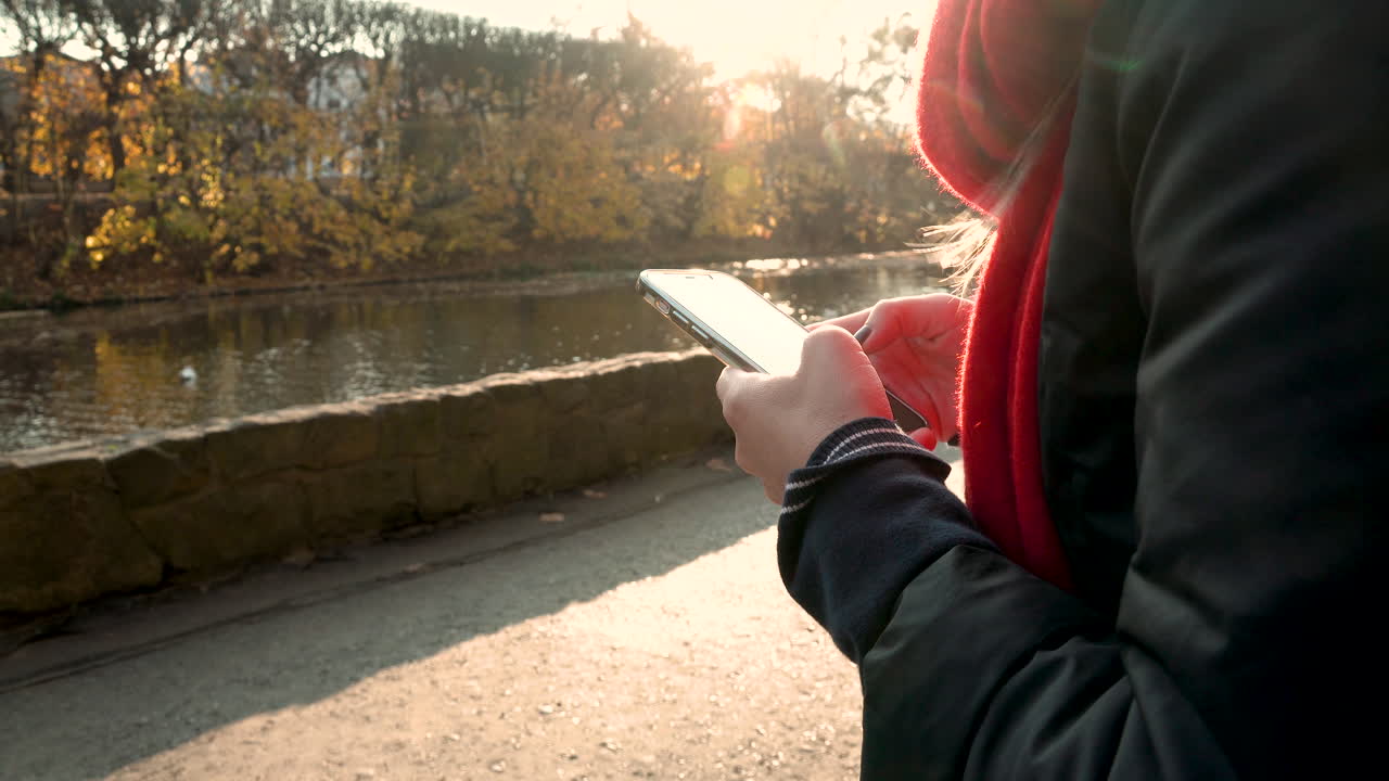 Close up of female person typing on smartphone outdoors beside river during sunset in background - 4K