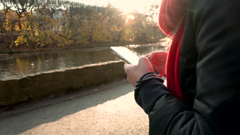 Close-up-of-female-person-typing-on-smartphone-outdoors-beside-river-during-sunset-in-background---4K