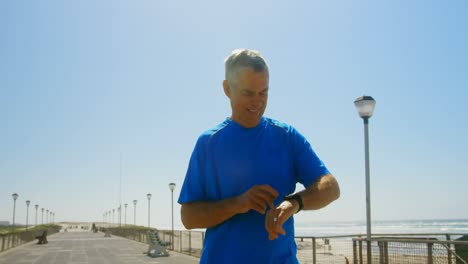 Vista-Frontal-De-Un-Hombre-Caucásico-Senior-Activo-Usando-Un-Reloj-Inteligente-En-Un-Paseo-Marítimo-En-La-Playa-4k