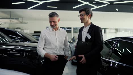 a confident guy a brunette assistant in glasses and a black body suit, communicates with a middle-aged man in a white shirt about a specific model of a modern white car in a car showroom