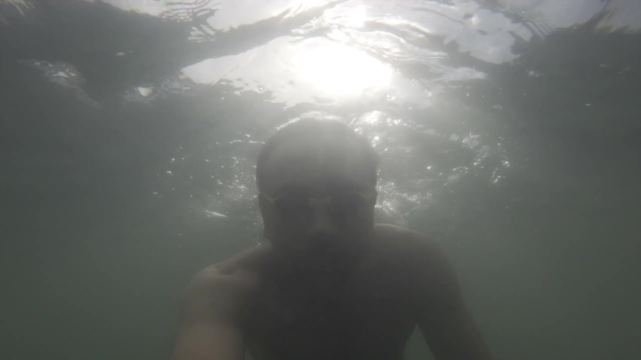 Hombre con gafas de natación nadando bajo el agua, tomándose una selfie, con el sol brillante brillando a través de la superficie del agua ondulante.