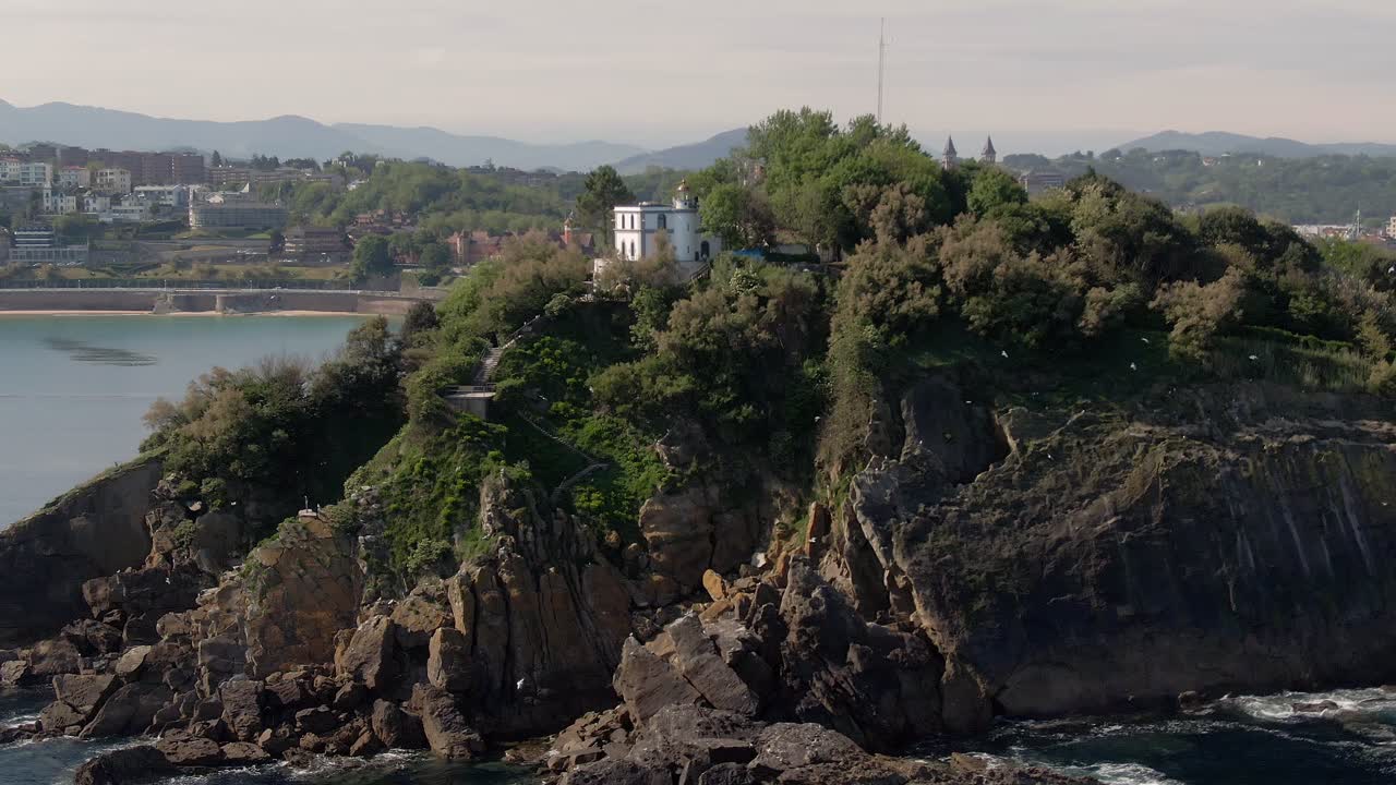 Solitario edificio bianco su un'isola rocciosa con uccelli in volo e San Sebastián sullo sfondo, vista aerea cinematografica