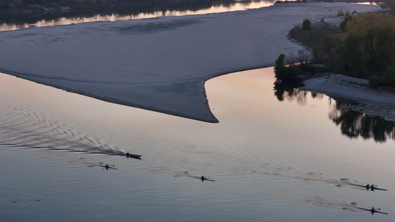 Synchronized rowers and a coach boat at sunset on the tranquil Po river near a sandy bank, with soft waves glowing in the fading light, reflecting the peaceful energy of early evening