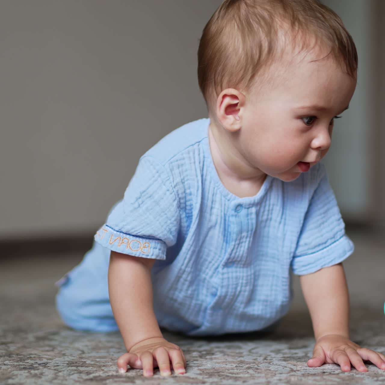 Nice cute baby boy in blue clothes stands on all fours on the floor. Lovely kid tries to choose a toy in front of him. Blurred backdrop
