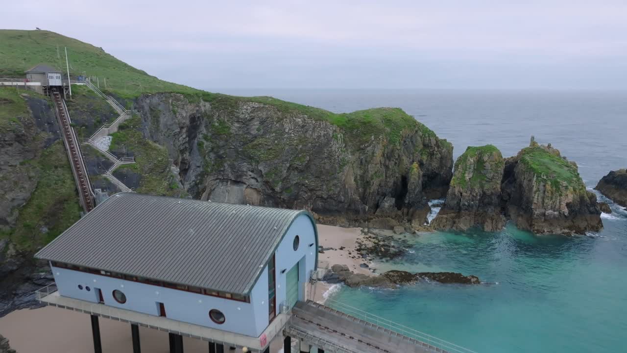 Lifeboat station with slipway descending into clear blue seawater next to jagged sea cliffs. Camera flight towards and over station. Mother Ivey's Bay, Cornwall, UK.