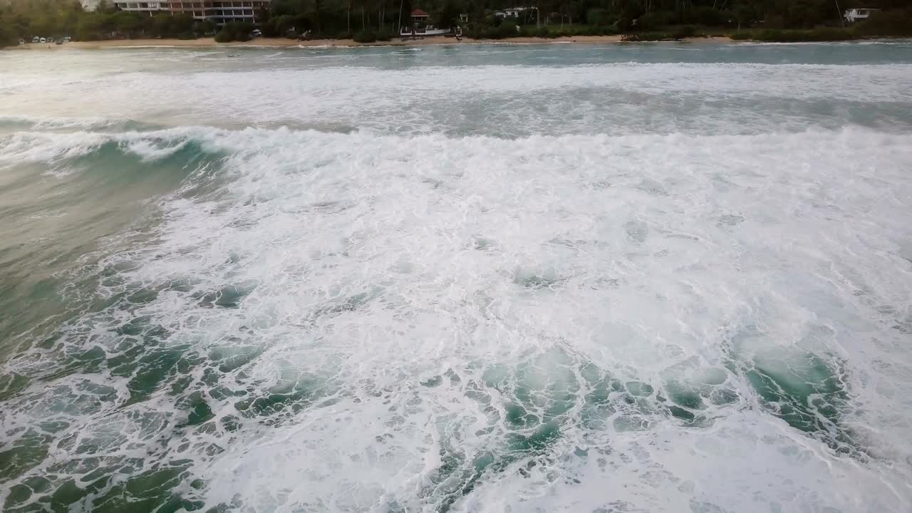 Drone flying backwards from peaceful ocean coastline revealing big foaming waves washing the shore on a sunny summer day