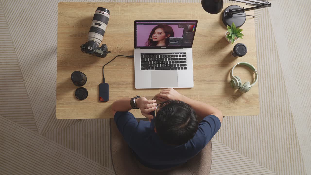 Top View Of A Male Editor Talking On Smartphone While Sitting In The Workspace Using A Laptop Next To The Camera Editing Photo Of A Female At Home