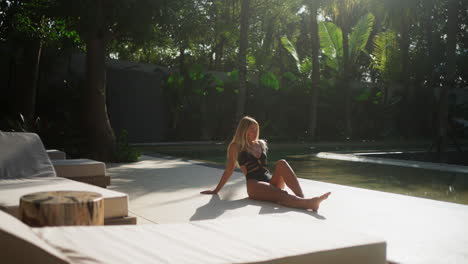 attractive, young blonde caucasian woman laying by luxury pool alone and sunbathing in tropical environment in black swimsuit in the sun - sliding shot, wide shot, slow motion.