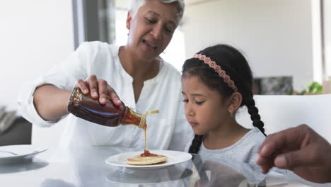 una abuela biracial vierte jarabe en un panqueque para una joven nieta biracial en una mesa de la cocina