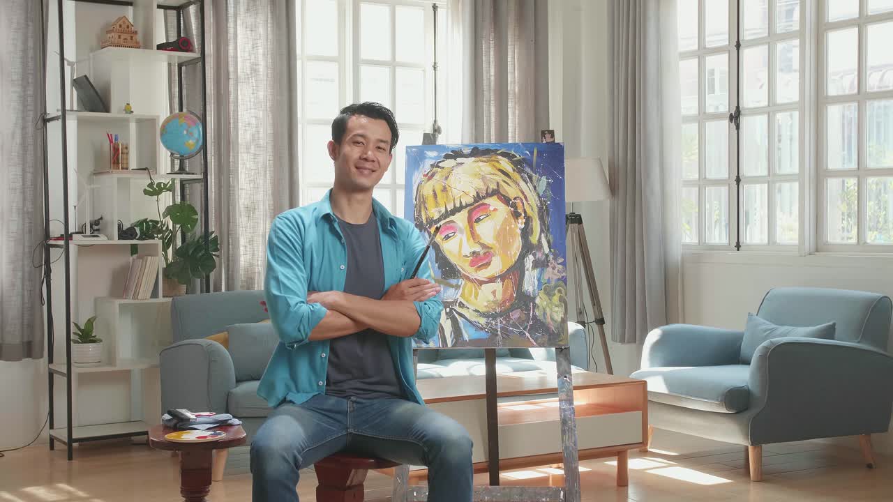Asian Artist Man Sitting On Round Wooden Chair Without Backrest Holding Paintbrush Mixed Colour, Crossing His Arms, And Smiling After Finish Painting A Girl On The Canvas