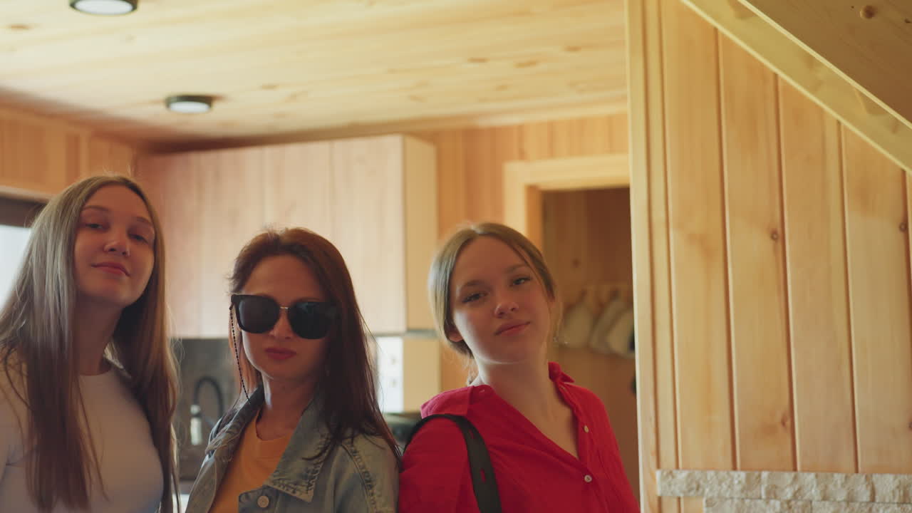 Three sisters stand side by side in wooden interior with warm lighting as middle sister lifts sunglasses with confident gesture while others pose casually