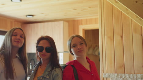 three sisters stand side by side in wooden interior with warm lighting as middle sister lifts sunglasses with confident gesture while others pose casually