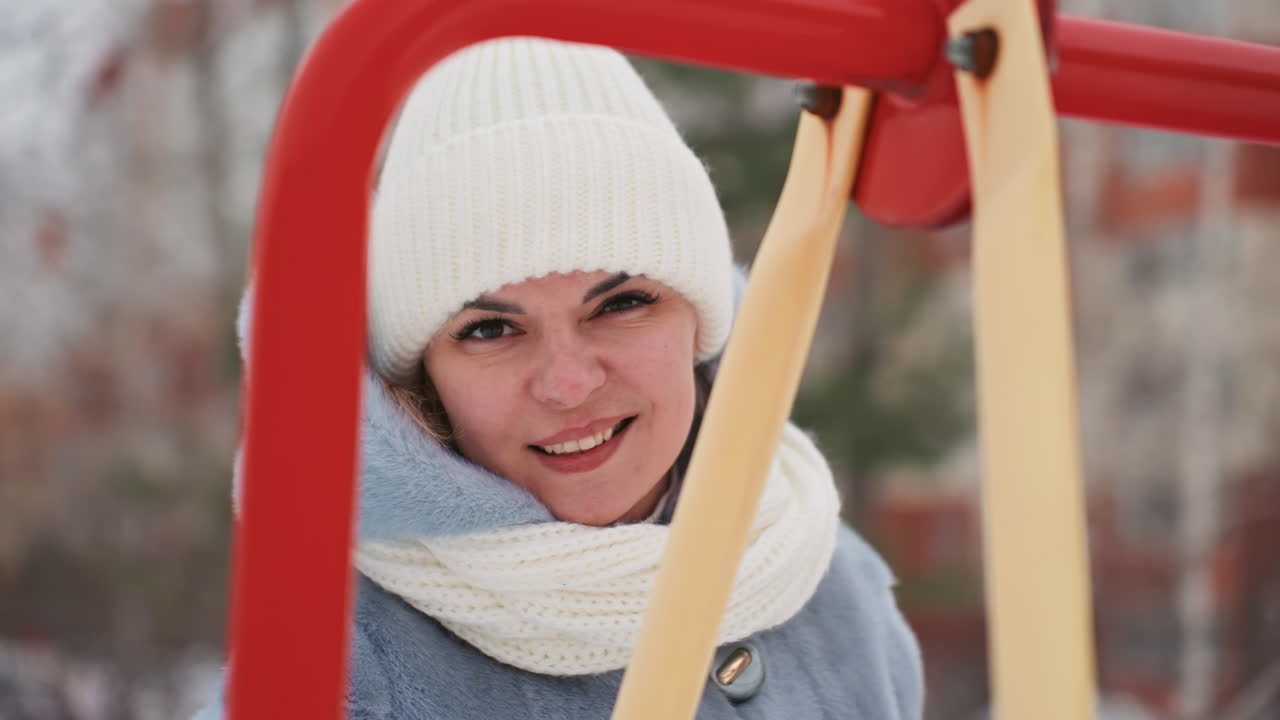 Mamma caucasica sorride sull'altalena innevata, gioca con il figlio nelle vicinanze, entrambi infagottati in berretto e sciarpa, espressione vivace e spontanea, risate nell'aria fredda, telaio rosso dell'altalena, profondità di campo ridotta, un gioioso momento familiare