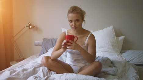 pretty woman in white tank top and panties sitting on bed and enjoying hot beverage